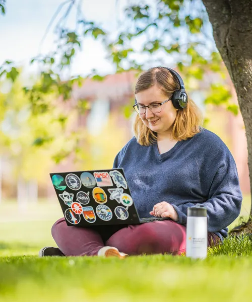 girl wearing headphones and working on a laptop while sitting under a tree